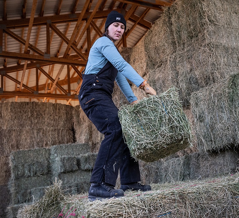 Image of a woman in Timberland overalls, blue long sleeve work t-shirt and black PRO beanie, standing on bales of hay in a barn, getting ready to throw a bale.