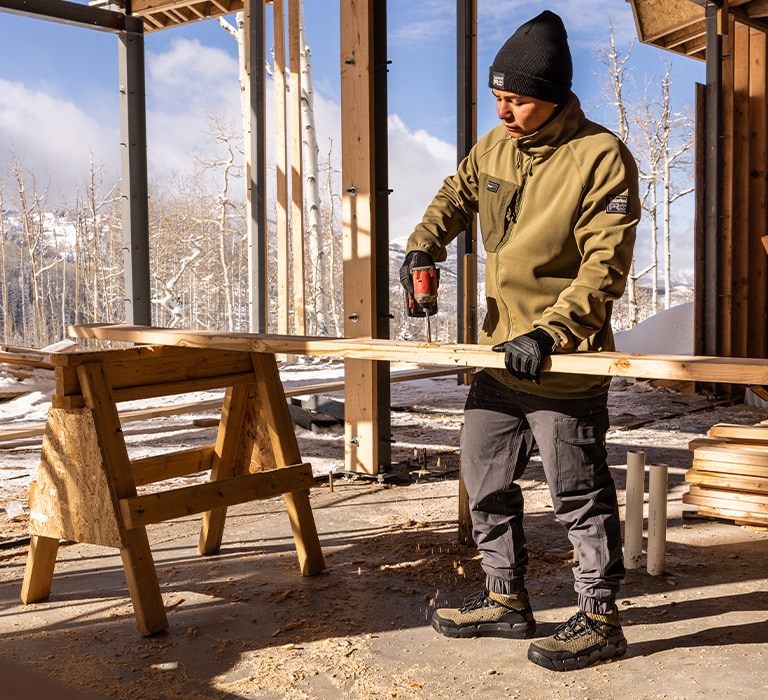 Image of a man standing in a framed open air construction site,  working on a piece of lumber on a sawhorse, wearing a black Timberland beanie, tan Timberland work hoodie, gray Morphix work jogger pants and brown Morphix work chukka shoes.