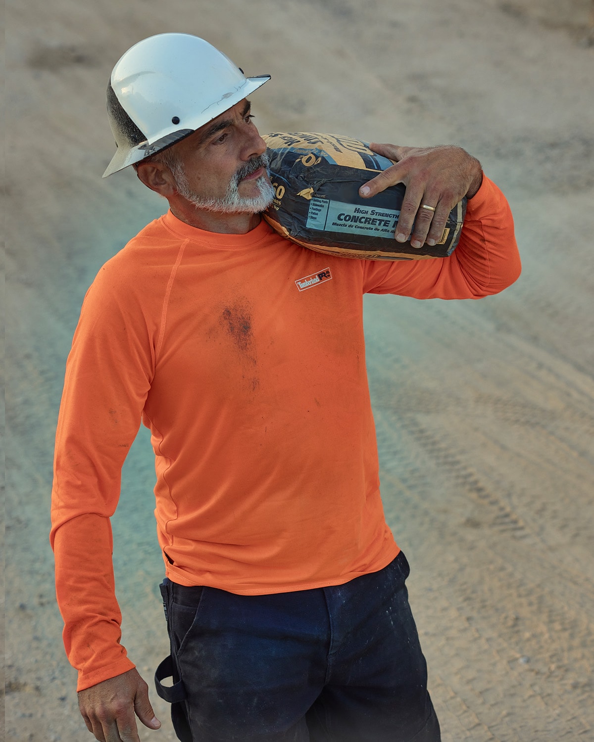 Image of a man in white hard hat, white cropped beard, orange Timberland PRO long sleeve t-shirt and dark blue work pants, walking in a sandy lot and carrying a bag of concrete on his shoulder.