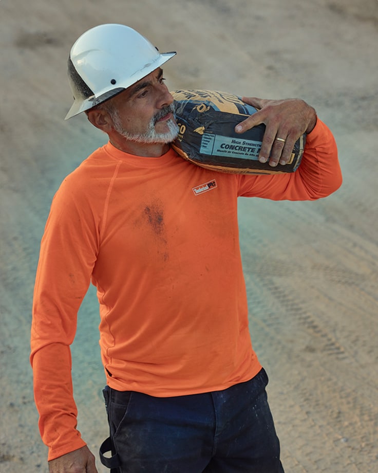 Image of a man in white hard hat, white cropped beard, orange Timberland PRO long sleeve t-shirt from the waist up, walking in a sandy lot and carrying a bag of concrete on his shoulder.
