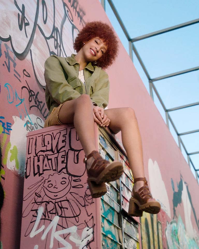 Image of a woman with short curly hair wearing a green jacket, shorts and brown strappy heeled Timberland sandals, smiling and hanging out in a graffiti-covered alleyway. Closeup image of a woman from the ankles down, wearing brown strappy heeled leather Timberland sandals.