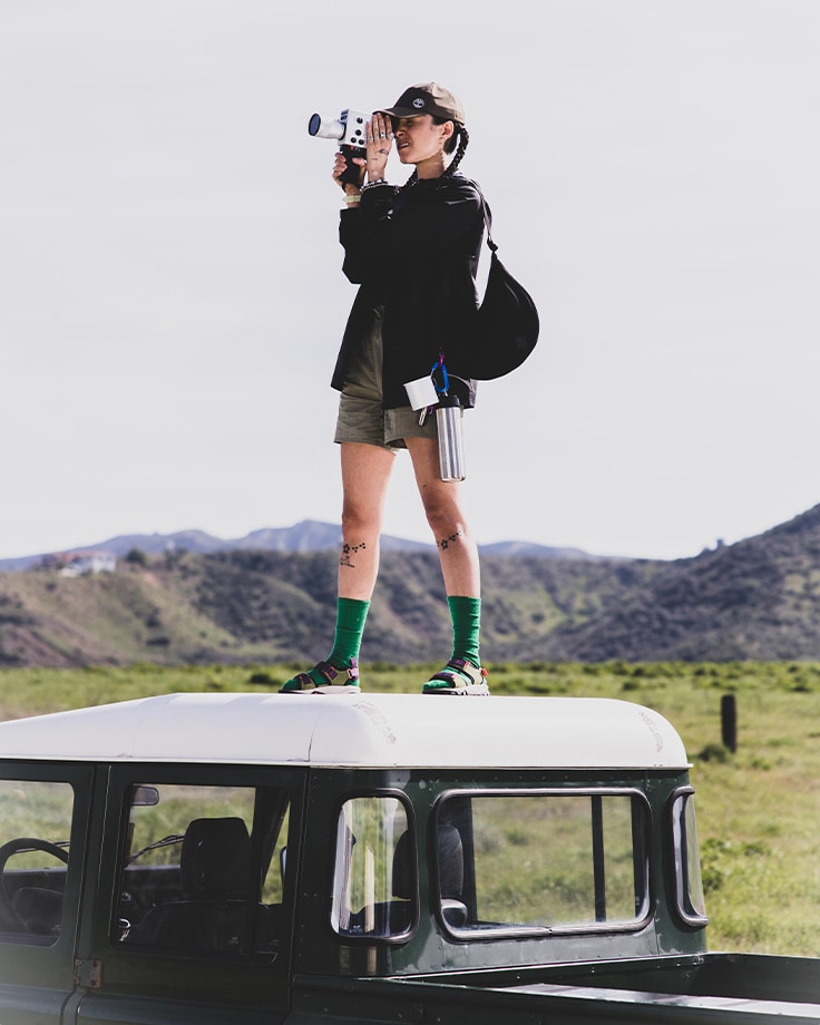 Image of a woman standing on the roof of a white car, in a green valley with mountains behind, looking through a camera lens and wearing black Timberland jacket, shorts, green socks and slide sandals.
