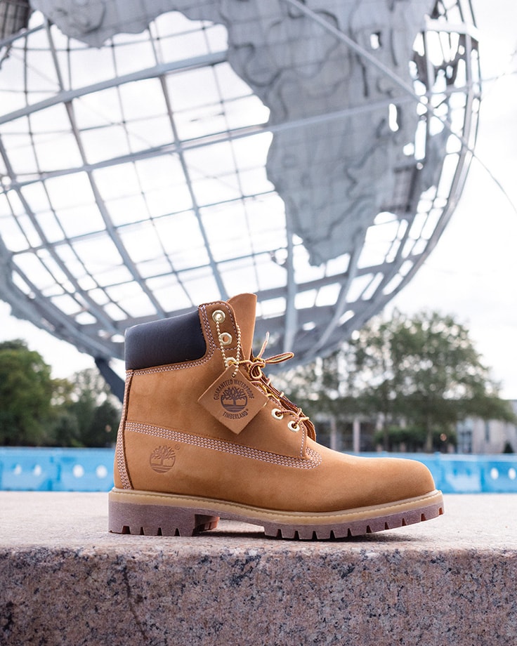 Image of the classic wheat Timberland boot on concrete with a large metal structure depicting the Earth and continents behind it.