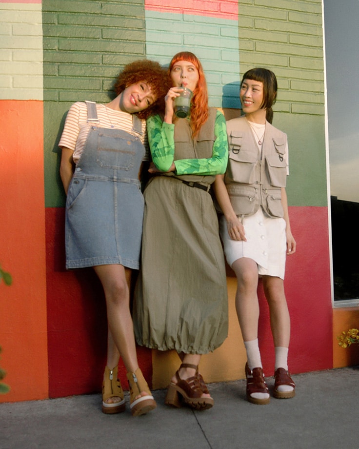 Image of three smiling young women standing against a colorblocked orange, red, green and white painted wall, all wearing Timberland leather strappy sandals and utility inspired workwear outfits.