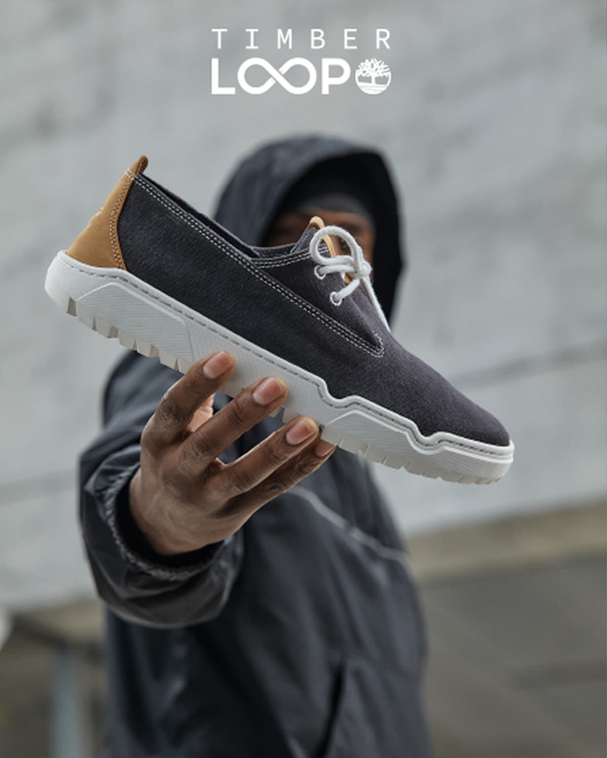 Image of a 2-eye black sneaker with white outsoles, being held up to the camera in sharp focus by a man in a hoodie in the background that's blurred. Image of a pair of navy 2-eye sneakers with white outsoles being held up by a man's hand.